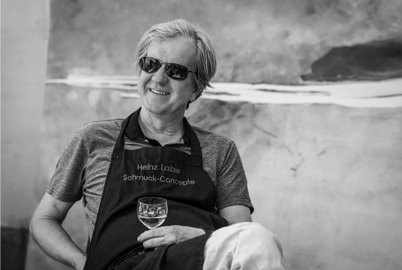 Ein Mann mit Sonnenbrille und einer Schürze, auf der Heinz Labile Schmuck-Concepte steht, sitzt entspannt lächelnd bei einem Glas Weißwein. Das Schwarz-Weiß-Bild hat einen abstrakten, unscharfen Hintergrund und fängt eine schicke Modehaus-Atmosphäre ein.